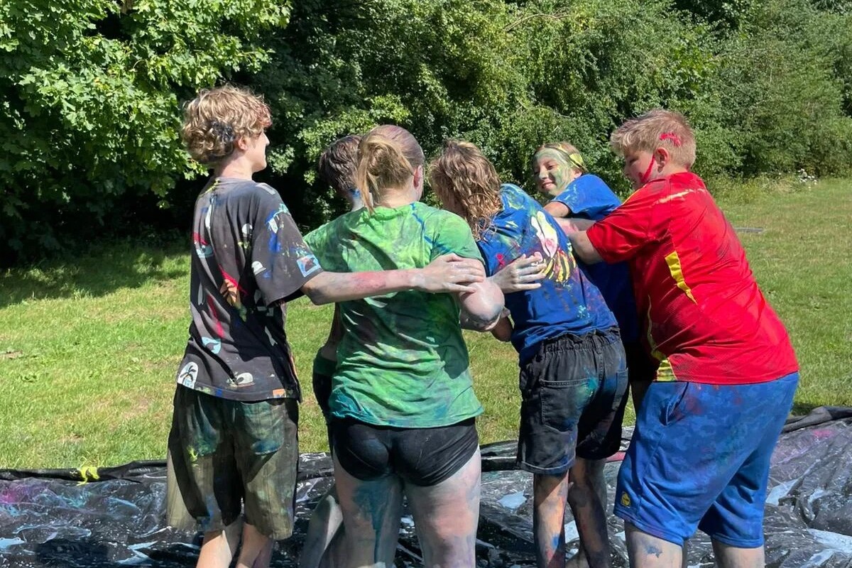 Zes mensen staan dicht bij elkaar op een met kleurrijke verf besmeurd zeil in de open lucht; hun kleding en benen zitten onder de vlekken. Ze staan met hun rug naar de camera en lijken te spelen of elkaar te omhelzen. Op de achtergrond staan groene bomen.