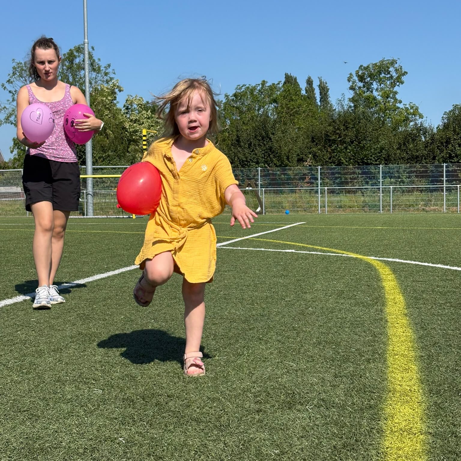 Een jong meisje in een gele jurk rent over een met gras begroeid sportveld met een rode bal in haar hand, terwijl achter haar een volwassene loopt die twee roze ballen bij zich heeft. De lucht is helder en blauw.