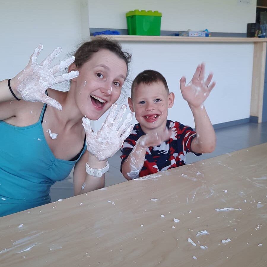 Een vrouw en een jongetje glimlachen en zwaaien naar de camera, terwijl ze hun handen laten zien die bedekt zijn met witte verf of plaksel. Ze zitten aan een tafel waarvan het oppervlak ook met wit plaksel is besmeurd.