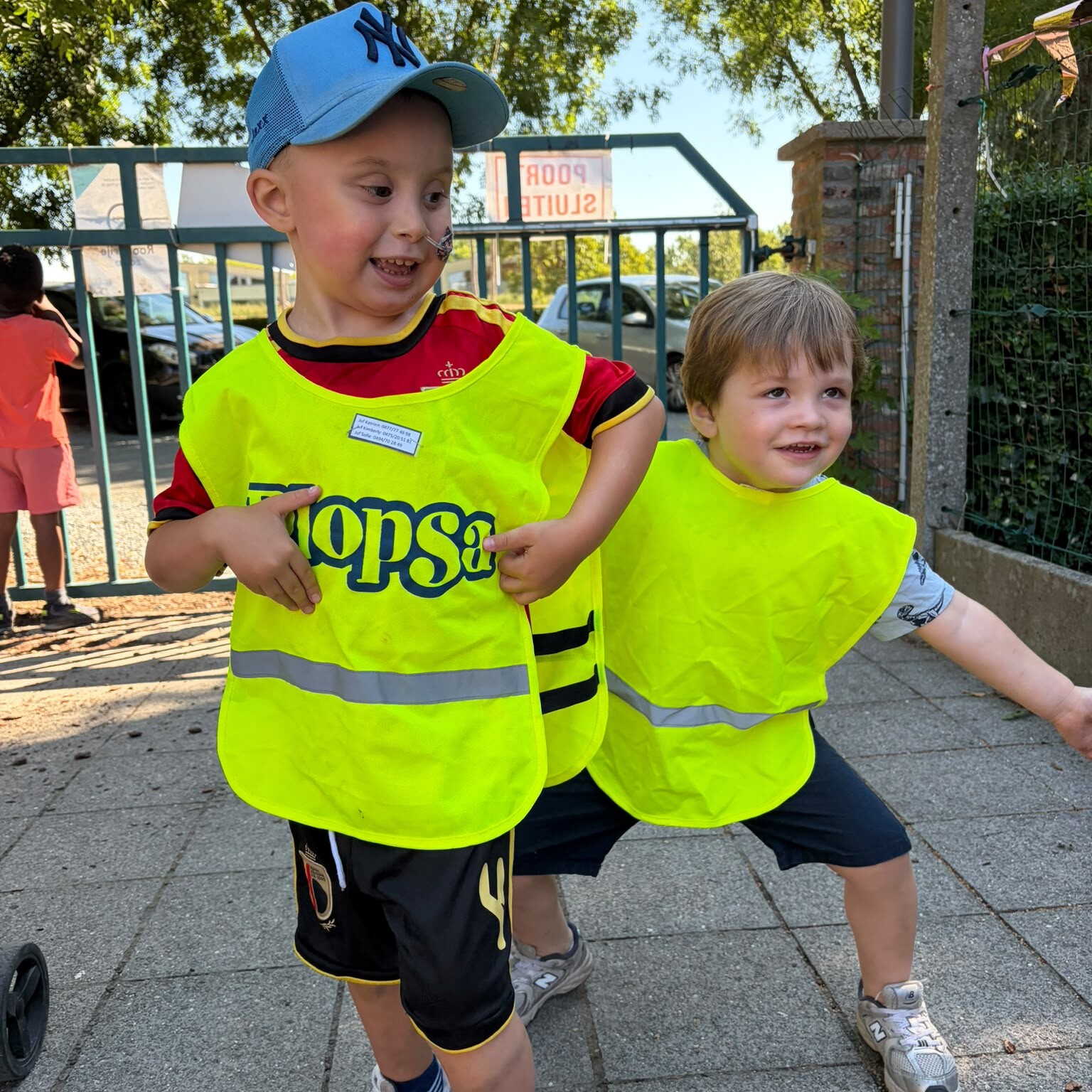 Twee jonge jongens in felgele veiligheidsvesten glimlachen en spelen samen buiten. De ene draagt een blauwe pet en een rood shirt; de andere draagt een grijs shirt. Op de achtergrond zijn een metalen hek, bomen en een bord te zien.