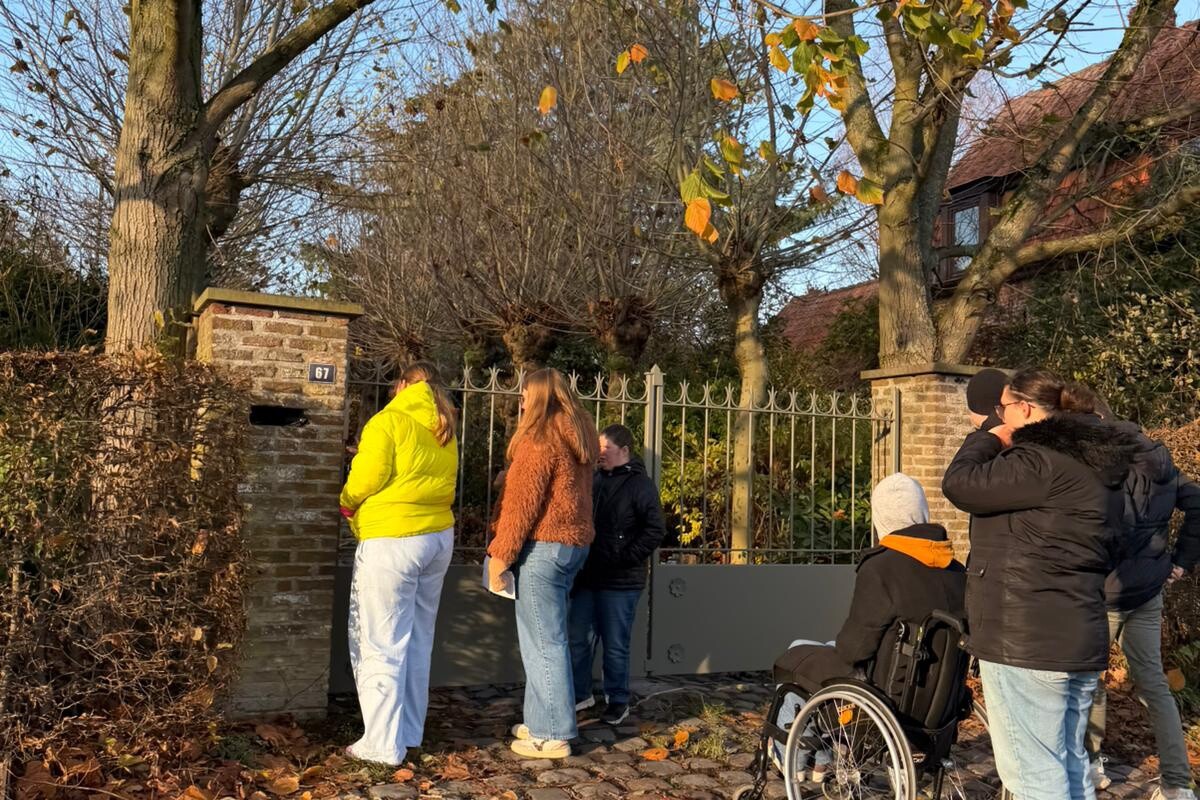 Vijf mensen, waaronder één in een rolstoel, staan op een geplaveid pad naast een poort en een bakstenen muur, omringd door kale bomen en gevallen herfstbladeren. Iedereen kijkt naar de poort en staart voor zich uit.