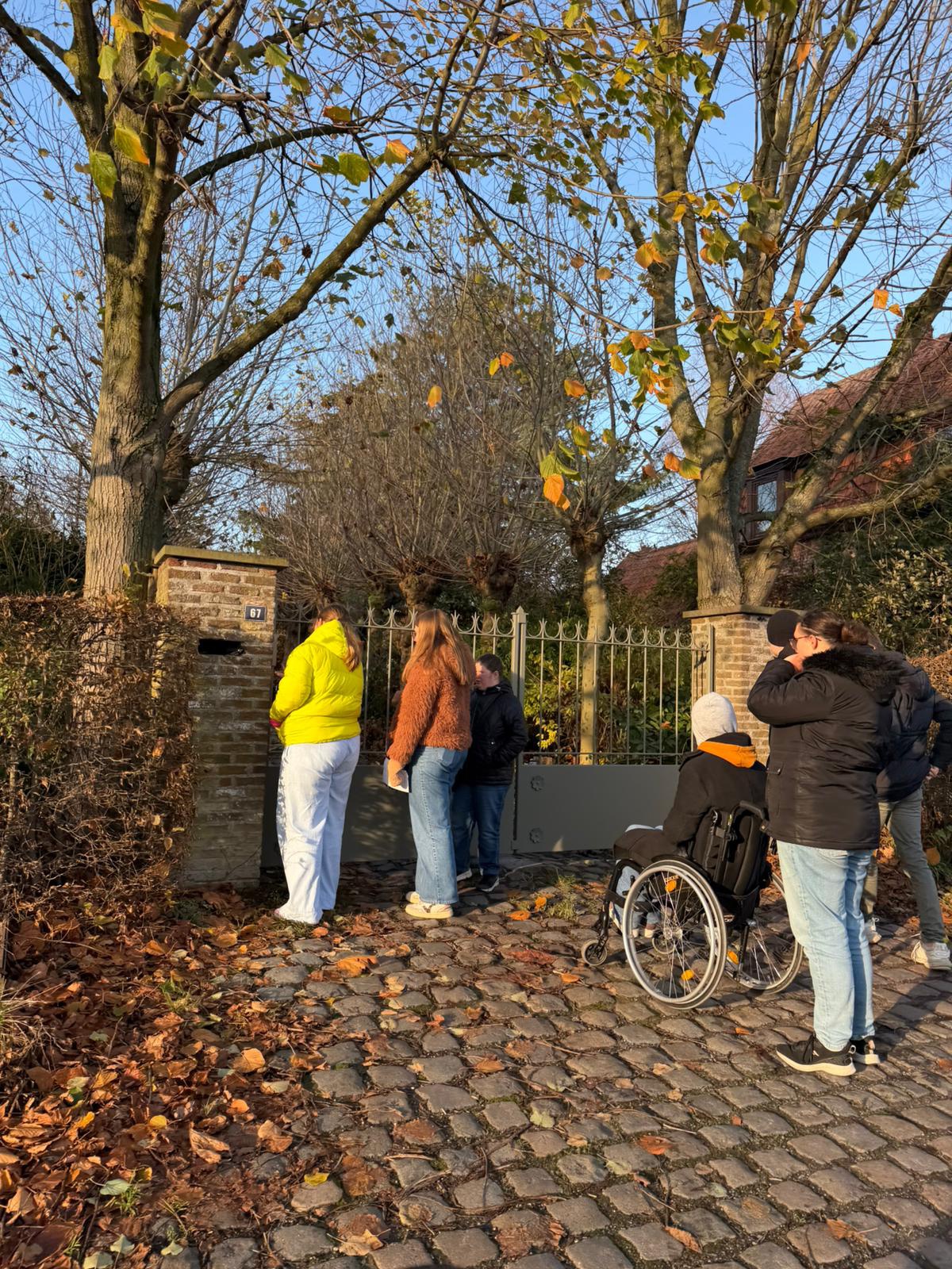 Vijf mensen, waaronder één in een rolstoel, staan op een geplaveid pad naast een poort en een bakstenen muur, omringd door kale bomen en gevallen herfstbladeren. Iedereen kijkt naar de poort en staart voor zich uit.